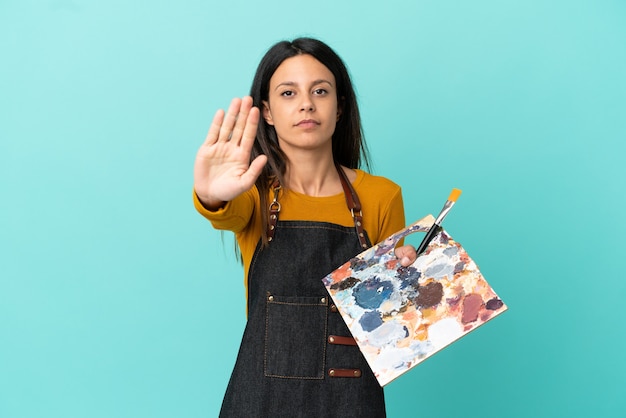 Joven artista mujer caucásica sosteniendo una paleta aislada sobre fondo azul haciendo gesto de parada