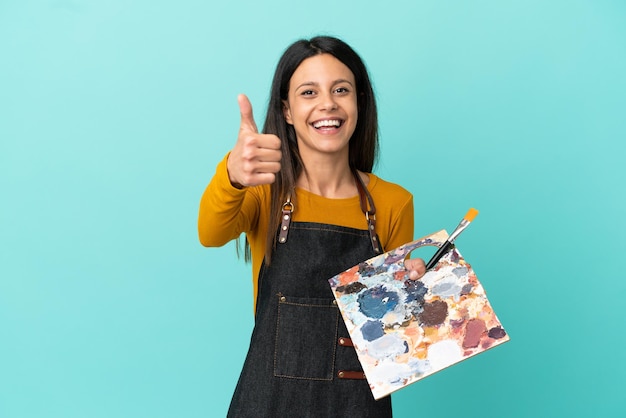 Joven artista mujer caucásica sosteniendo una paleta aislada sobre fondo azul con los pulgares hacia arriba porque algo bueno ha sucedido