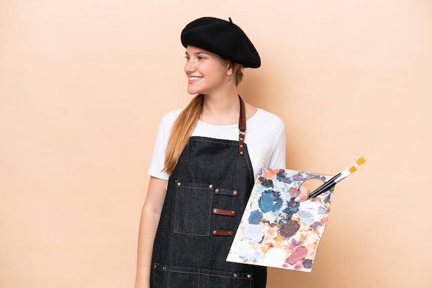 Joven artista mujer caucásica sosteniendo una paleta aislada sobre fondo beige mirando hacia el lado