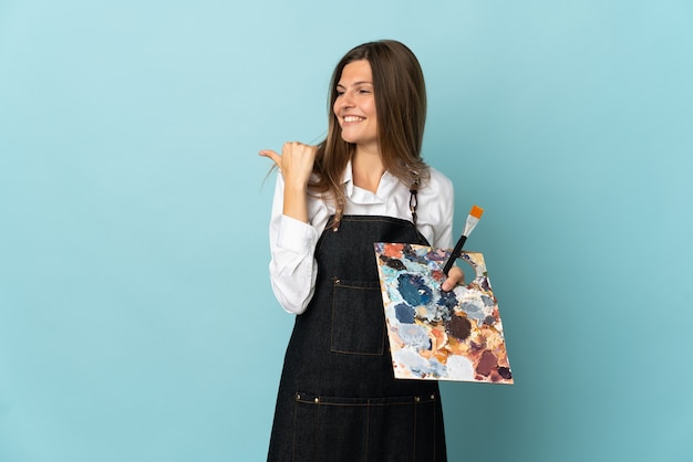 Joven artista mujer eslovaca aislada sobre fondo azul apuntando hacia el lado para presentar un producto