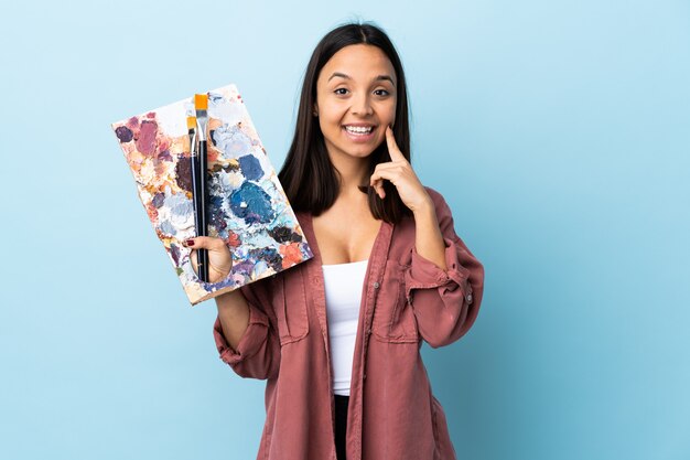 Joven artista mujer sosteniendo una paleta sobre azul aislado sonriendo con una expresión feliz y agradable