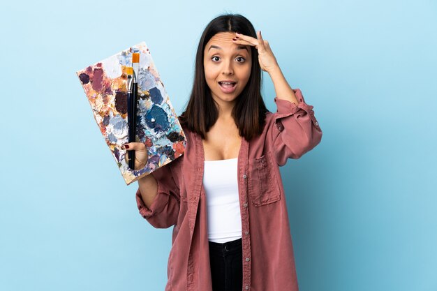 Joven artista mujer sosteniendo una paleta sobre pared azul aislado saludando con la mano con expresión feliz