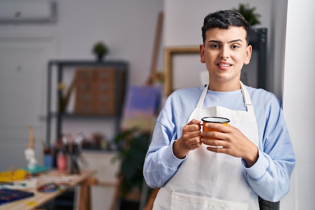 Joven artista no binario sonriendo confiado bebiendo café en el estudio de arte