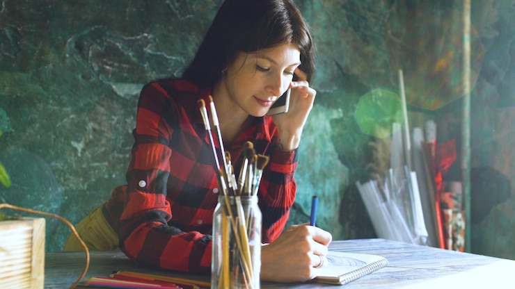 Foto joven artista pintando scetch en un cuaderno de papel con lápiz y hablando por teléfono en el interior