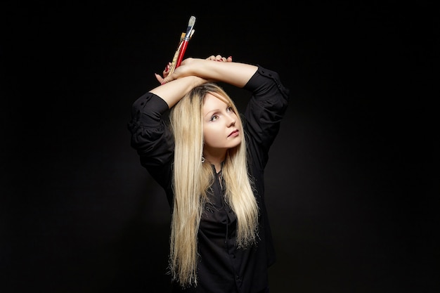 Joven artista rubia posa con un pincel en la mano. sesión de fotos sobre un fondo negro en el estudio.