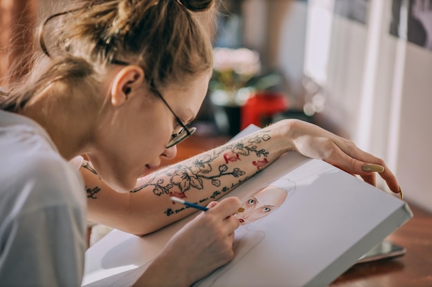 Joven artista se sienta a la mesa y pinta el retrato de una mujer sobre lienzo con pinturas de acuarela Dibujar como hobby Escena real