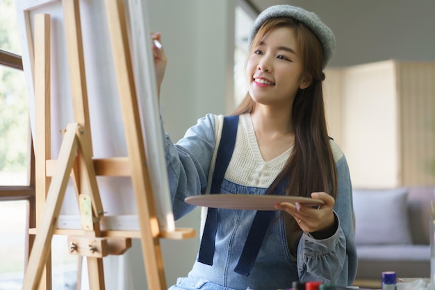 La joven artista sostiene una paleta de colores y usa un pincel para pintar obras de arte sobre lienzo en un estudio en casa