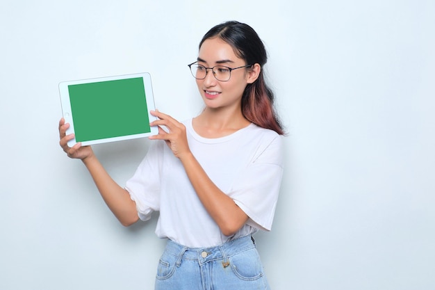 Una joven asiática alegre con una camiseta blanca que muestra una tableta digital con una pantalla vacía aislada de fondo blanco