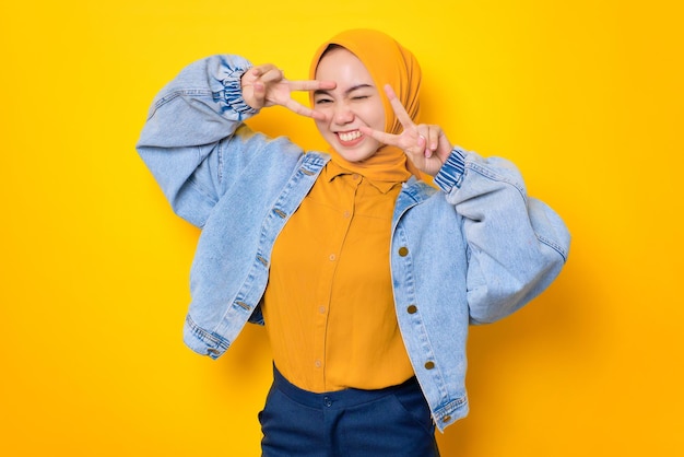 Una joven asiática alegre con una chaqueta de jeans que muestra el signo de la paz sobre los ojos aislados sobre un fondo amarillo