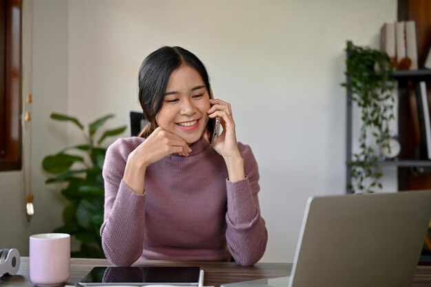 Una joven asiática alegre haciendo una llamada telefónica con su amiga en su mesa de estudio