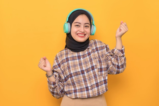 Una joven asiática alegre con pantalones a cuadros escuchando su música favorita con auriculares y bailando aislada de fondo amarillo