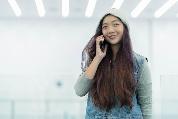 Joven asiática alegre con ropa informal que tiene una conversación telefónica parada en un centro comercial, una mujer independiente sonriente que llama a un colega para compartir buenas noticias sobre negocios