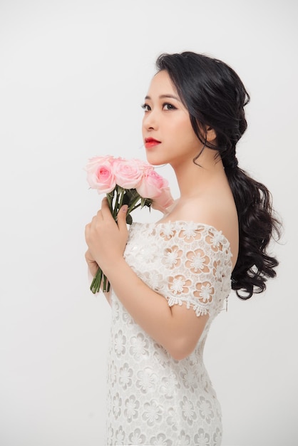 Una joven asiática alegre con vestido blanco y sosteniendo un ramo de flores sobre fondo blanco