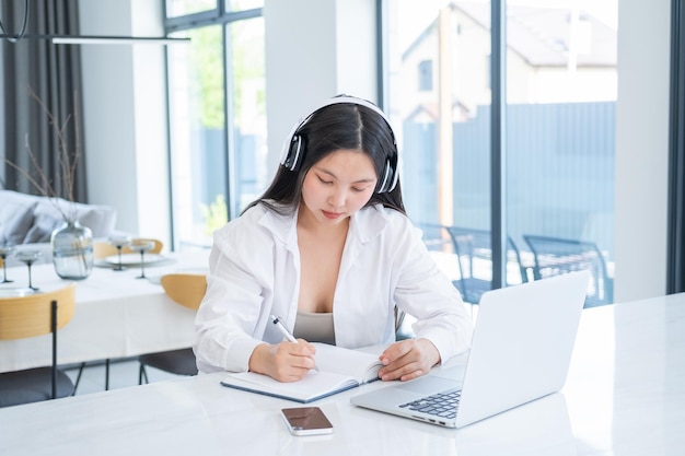 Joven asiática con auriculares inalámbricos haciendo notas en un libro diario con una computadora portátil abierta en una casa moderna