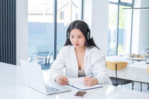 Joven asiática con auriculares inalámbricos haciendo notas en un libro diario con una computadora portátil abierta en una casa moderna