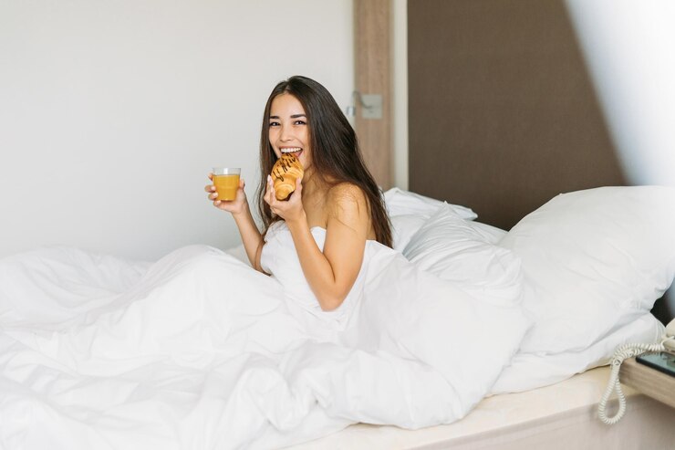 Foto joven asiática con cabello largo desayunando croissant y jugo de naranja en la cama de la habitación del hotel