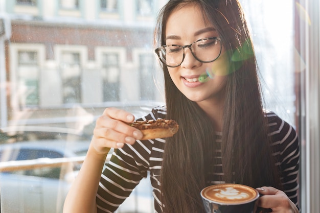 Joven asiática comiendo pastel en el alféizar de la ventana