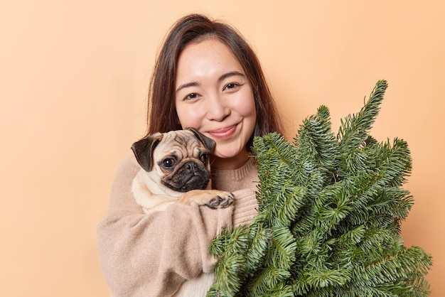 Una joven asiática complacida abraza con amor a su perro favorito, posa con ramas de abeto verde, sonríe tiernamente a la cámara y disfruta de una acogedora atmósfera doméstica, una mascota amante aislada sobre un fondo marrón