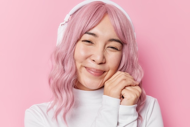 Una joven asiática complacida sonríe suavemente, mantiene las manos cerca de la cara, tiene el pelo teñido de rosa, escucha su música favorita a través de auriculares inalámbricos vestidos con un jersey blanco casual aislado sobre una pared rosada del estudio