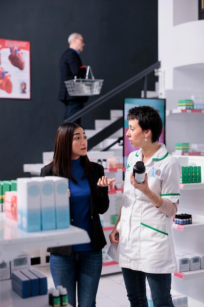 Joven asiática comprando vitamina y consultando con un trabajador de farmacia. Empleado de una tienda minorista médica que sostiene una botella de píldoras multivitamínicas y responde a las preguntas de los clientes sobre los aditivos alimentarios