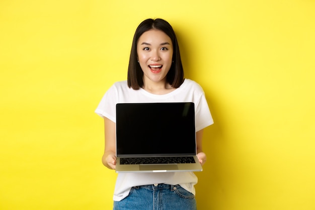 Joven asiática demuestra oferta en línea, mostrando la pantalla del portátil en blanco y sonriendo, de pie sobre fondo amarillo