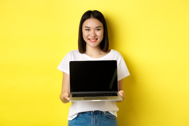 Joven asiática demuestra oferta en línea, mostrando la pantalla del portátil y sonriendo, de pie sobre fondo amarillo.