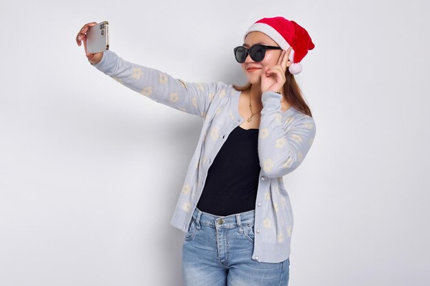 Una joven asiática emocionada con un sombrero de Navidad haciendo una selfie en un teléfono móvil publica una foto en una red social aislada de fondo blanco