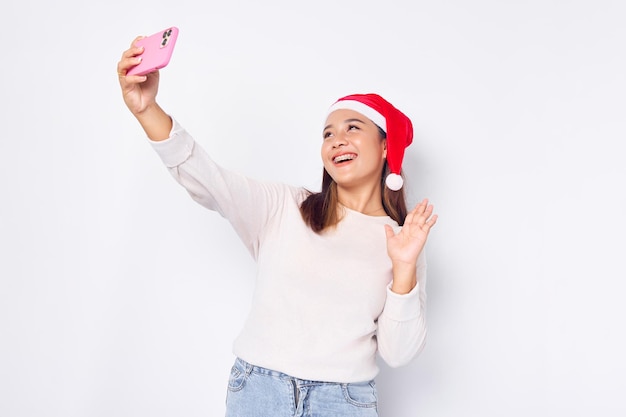 Una joven asiática emocionada con un sombrero de Navidad haciendo una selfie en un teléfono móvil publica una foto en una red social aislada de fondo blanco