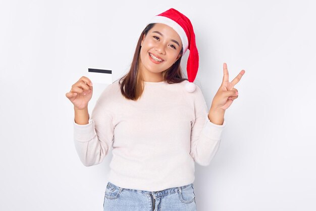 Una joven asiática emocionada con un sombrero de Navidad que muestra una tarjeta de crédito blanca en blanco que muestra el signo de la paz con el dedo aislado en el fondo blanco