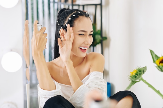 Joven asiática entrecerrar los ojos y cubrirse la cara con la mano al aplicar laca para el cabello