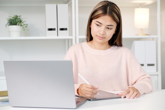 Joven asiática escribiendo en una tableta digital mientras mira una computadora portátil