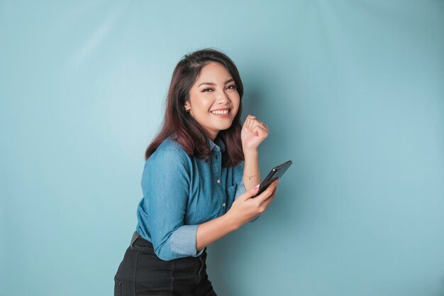 Una joven asiática con una expresión feliz y exitosa con un pantalones azul sosteniendo un teléfono inteligente aislado de fondo azul