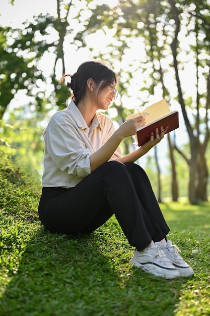 Una joven asiática feliz disfrutando leyendo un libro bajo el árbol en el parque verde