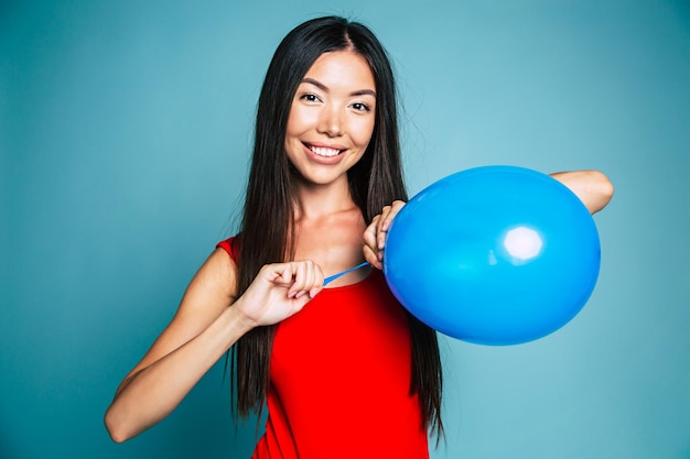 Una joven asiática feliz y emocional vestida de rojo está soplando un globo azul en el estudio. Retrato de una hermosa niña china. concepto de compras