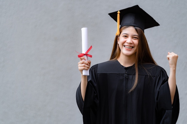 Una joven asiática feliz graduada universitaria en toga de graduación y birrete tiene un certificado de grado que celebra los logros educativos en el campus universitario. Educación foto de stock