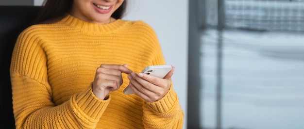 Una joven asiática feliz se relaja en un cómodo escritorio en casa enviando mensajes de texto en su teléfono inteligente una niña sonriente usa el teléfono celular hablando en línea mensajería compras en línea desde casa