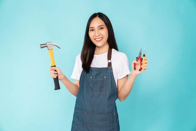 Foto una joven asiática feliz usa un delantal y sostiene un martillo de carpintero con un destornillador de pinzas en el fondo azul