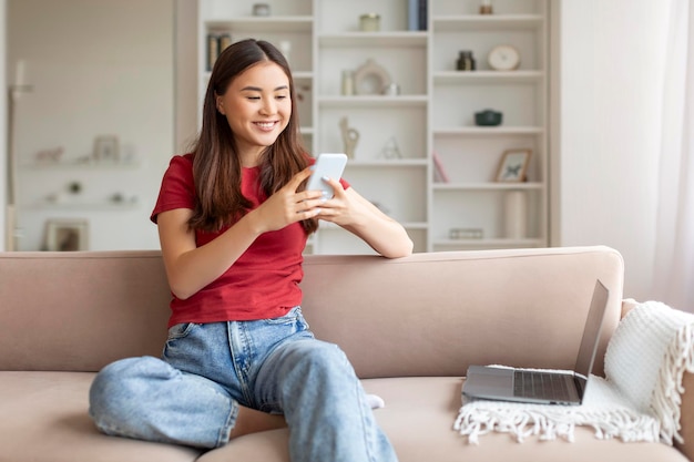 Una joven asiática feliz usando un teléfono inteligente y una computadora portátil en casa