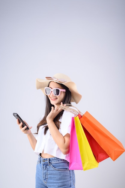 Foto joven asiática con gafas y sombrero lleva bolsas de la compra mientras juega smartphone sobre fondo blanco.