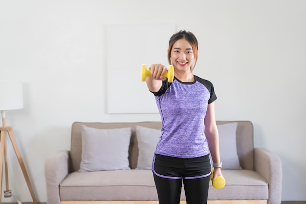 Joven asiática haciendo ejercicio y levantando pesas para entrenar brazos fuertes en casa
