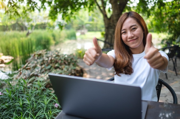 Una joven asiática haciendo y mostrando los pulgares hacia arriba mientras trabaja en una computadora portátil al aire libre