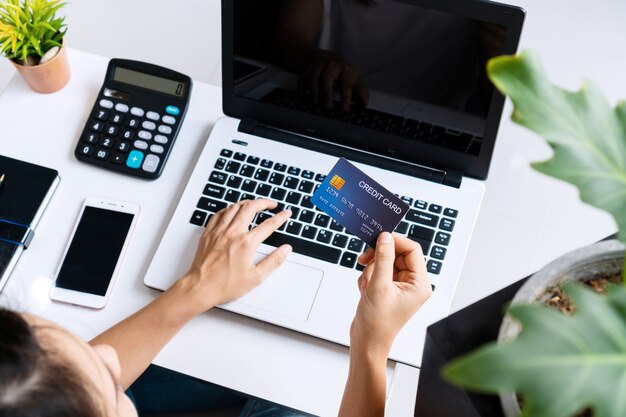 Foto joven asiática haciendo trabajos de investigación para su negocio con computadora portátil mientras sostiene la tarjeta de crédito en casa, vista superior y espacio de copia