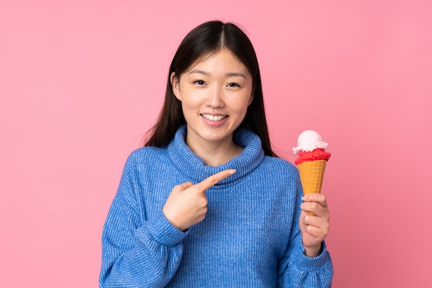 Joven asiática con un helado de cucurucho en la pared rosa y apuntando