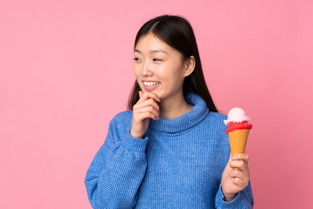 Joven asiática con un helado de cucurucho en la pared rosa pensando en una idea y mirando hacia el lado