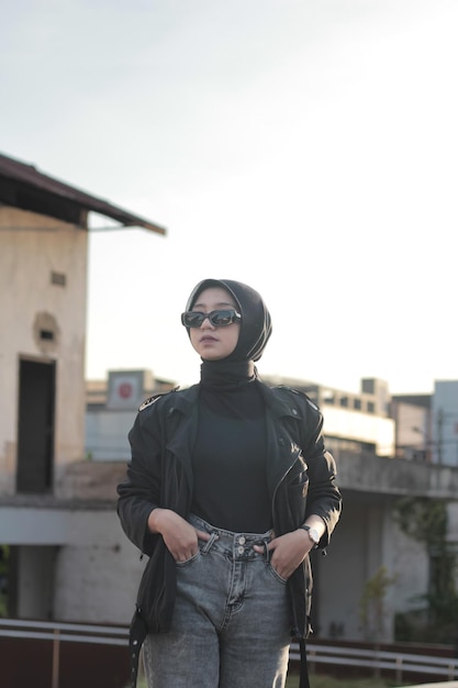 Una joven asiática con hijab y vaqueros posando al aire libre siguiendo un concepto con un outf negro