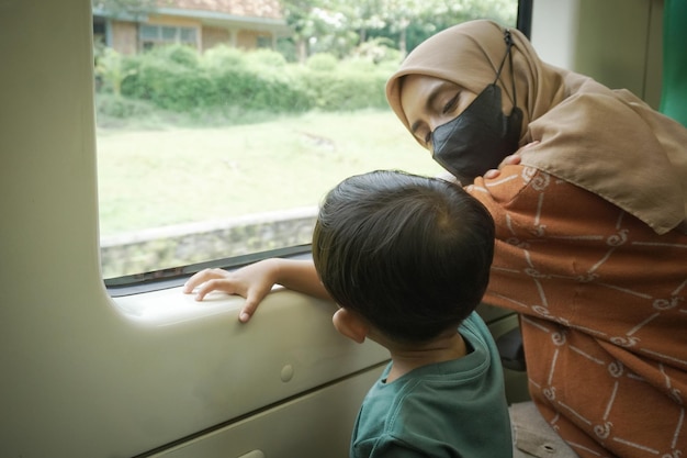 Una joven asiática con hiyab y su hijo están hablando y mirando por la ventana dentro del tren