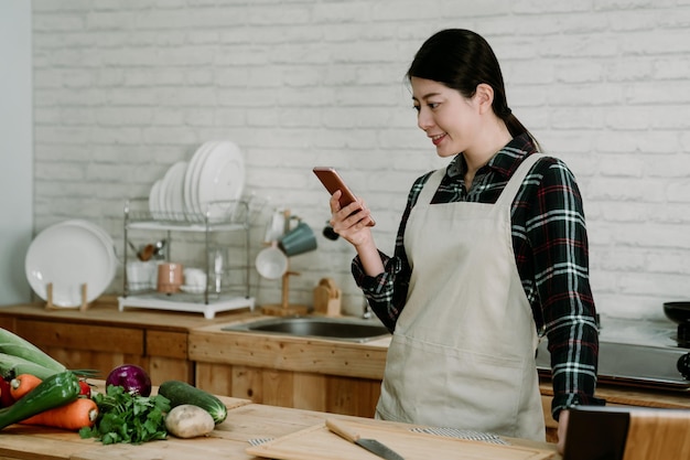 Joven asiática japonesa con verduras frescas sonriendo mientras usa el teléfono móvil en la cocina moderna. hermosa dama en mensaje de texto de delantal en línea en el teléfono celular durante el tiempo de cocción con comida saludable.