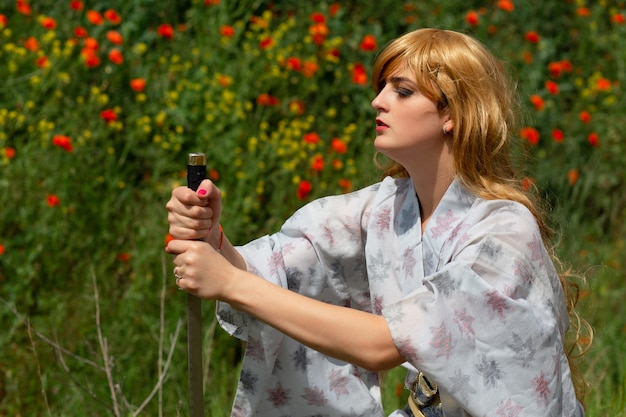 Joven asiática en kimono tradicional entrena técnicas de lucha con espada katana en las colinas con amapolas rojas, chica guerrera samurai