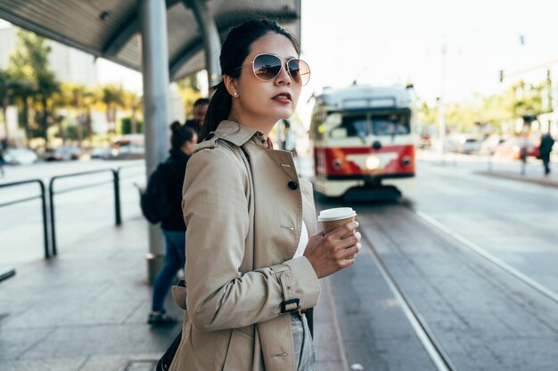 Joven asiática local con gafas de sol mirando alrededor sosteniendo una taza de café esperando el autobús en la estación. conmutar el concepto de estilo de vida activo. llegó el teleférico streeet car buceo en ferrocarril.