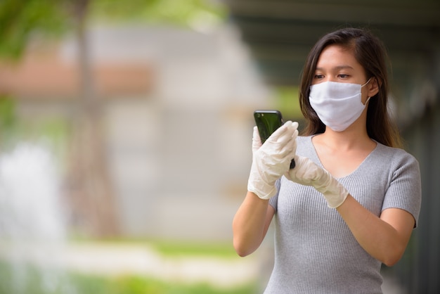 Joven asiática con máscara y guantes mientras usa el teléfono al aire libre